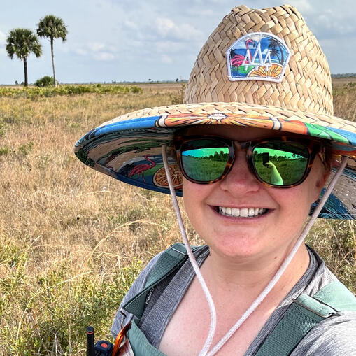 Shannon at UF/IFAS DeLuca Preserve in Okeechobee, FL. Shannon stands in an old field in central florida, wearing a straw hat and sunglasses. She smiles as the sun shines over the wild landscape.